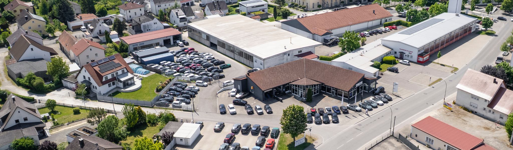 Luftaufnahme eines Autohauses mit geparkten Autos, Räder & Reifen, nahe gelegenen Gebäuden und Bäumen.
