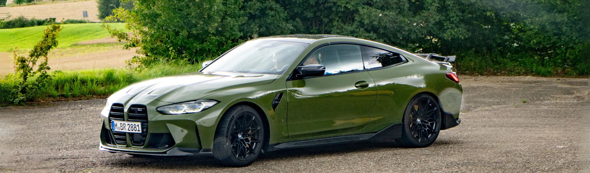 Ein grüner BMW parkt auf einer gepflasterten Fläche in der Nähe von Gras und Bäumen.