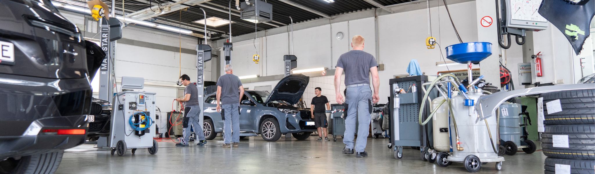 In einer geräumigen, gut beleuchteten Autowerkstatt arbeiten Mechaniker an Autos und bearbeiten Räder & Reifen.