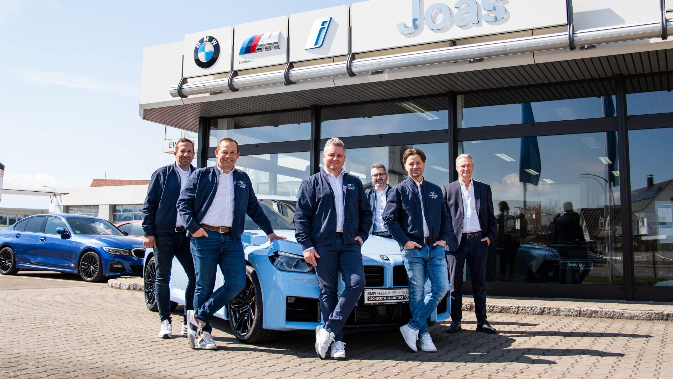 Sechs Männer posieren mit einem blauen Auto vor einem BMW-Autohaus.