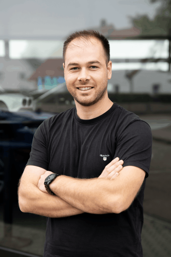 Ein Teammitglied in einem schwarzen T-Shirt steht draußen mit verschränkten Armen und lächelt in die Kamera.