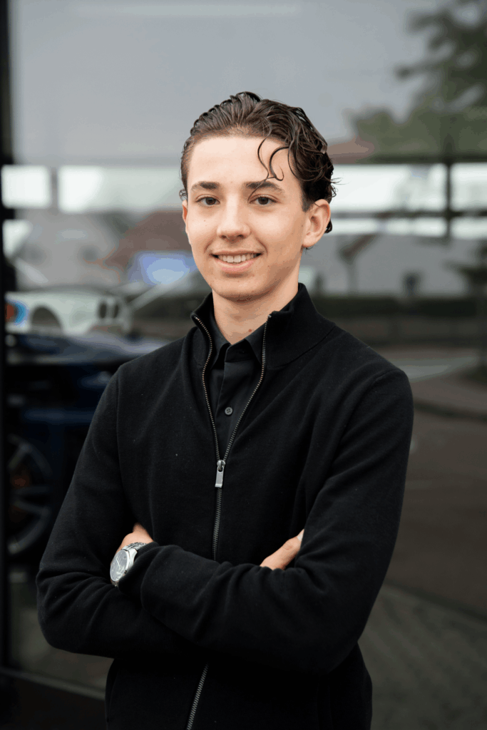 Ein junges Teammitglied in einer schwarzen Jacke steht draußen mit verschränkten Armen und lächelt in die Kamera.