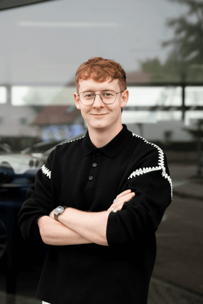 Ein junges Teammitglied mit Brille und roten Haaren steht im Freien, die Arme verschränkt und lächelt.