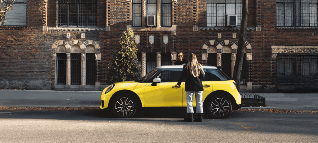 Zwei Frauen stehen an einem gelben Auto, das auf einer Straße in der Stadt vor einem Backsteingebäude geparkt ist.