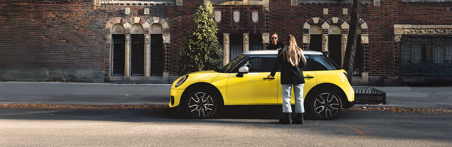 Zwei Personen stehen an einem gelben Jahreswagen, der auf einer Straße vor einem Backsteinhaus geparkt ist.