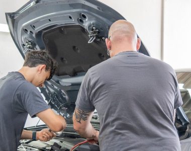Zwei Mechaniker inspizieren in einer hell erleuchteten Autowerkstatt Räder & Reifen unter der Motorhaube.